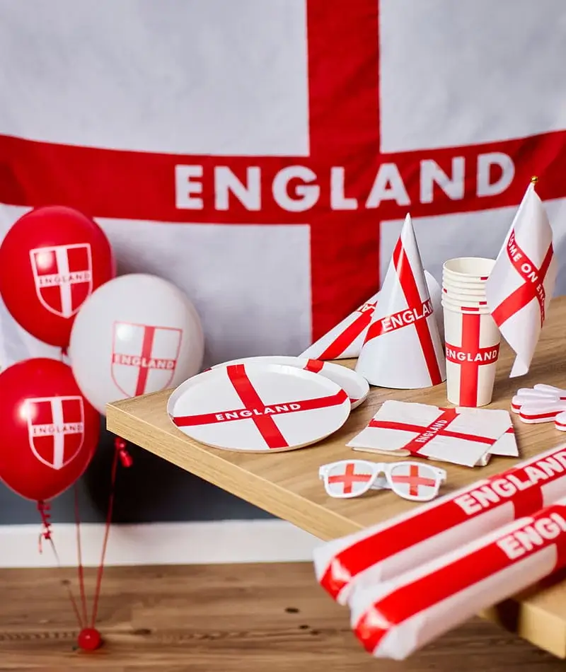 An England-themed party setup featuring red and white balloons, flags, paper plates, cups, napkins, and inflatable clapper sticks arranged on a wooden table in front of an England flag backdrop.